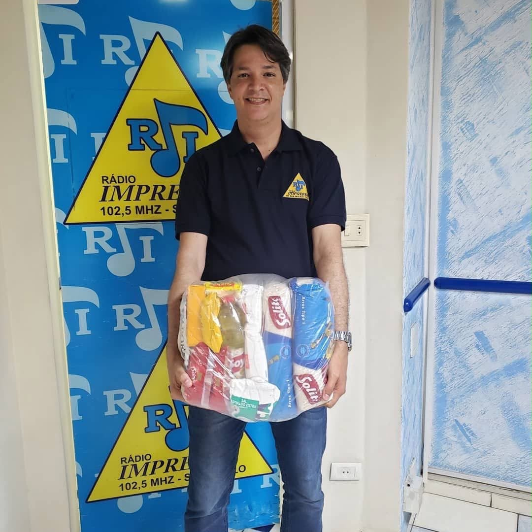 Gil do Povo com cesta de alimentos em ação solidária, em frente ao painel da Rádio Imprensa 102,5 MHz